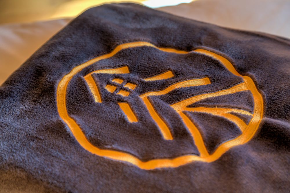 Close-up of a dark towel with an orange embroidered logo featuring a house and a road inside a circle.