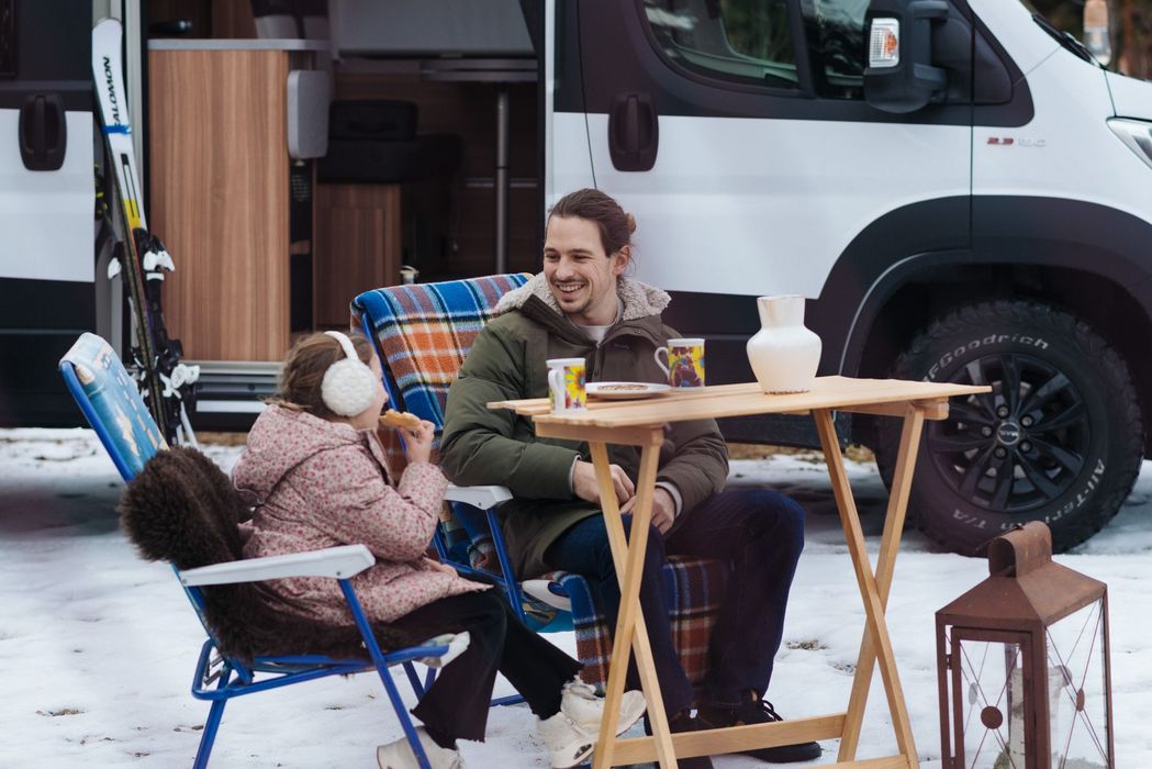 Padre e figlia seduti all'aperto sulla neve vicino a un camper, mentre mangiano e sorridono.
