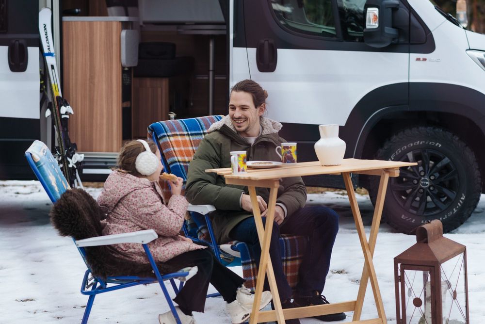 Mann und Kind sitzen im Winter vor einem Camper und essen Kekse.