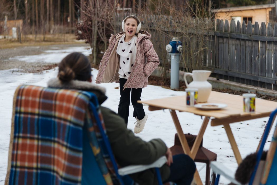 Bambina sorridente con giacca invernale e paraorecchie che corre sulla neve verso una persona seduta vicino a un tavolo.