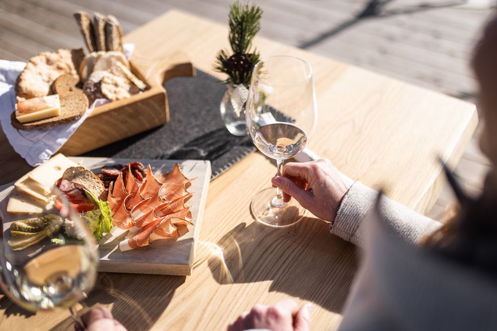 Person hält ein Weinglas neben einer Platte mit Aufschnitt, Käse und Brot auf einem Holztisch.