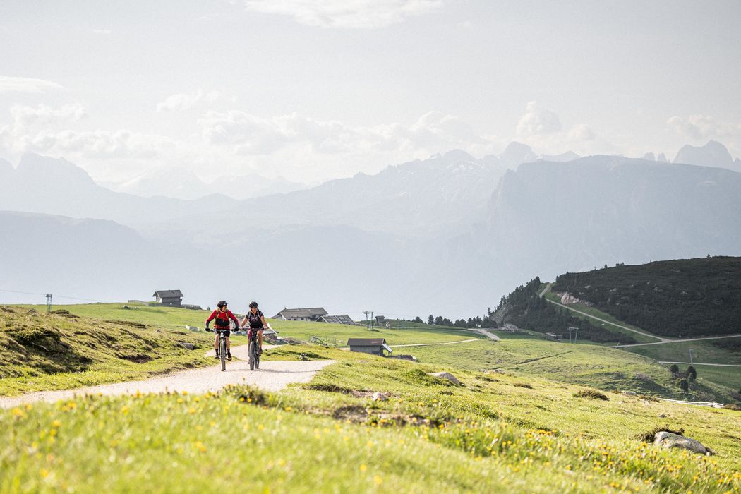 Due ciclisti su una strada di montagna verde con case e montagne sullo sfondo
