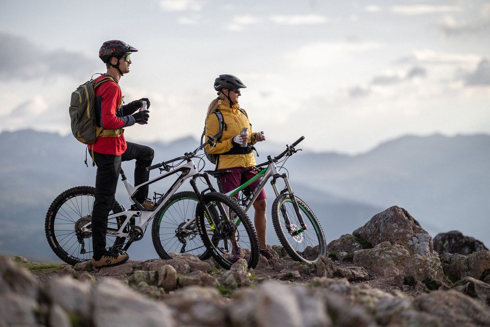 Two mountain bikers taking a break on rocky terrain with bikes, drinking from bottles.