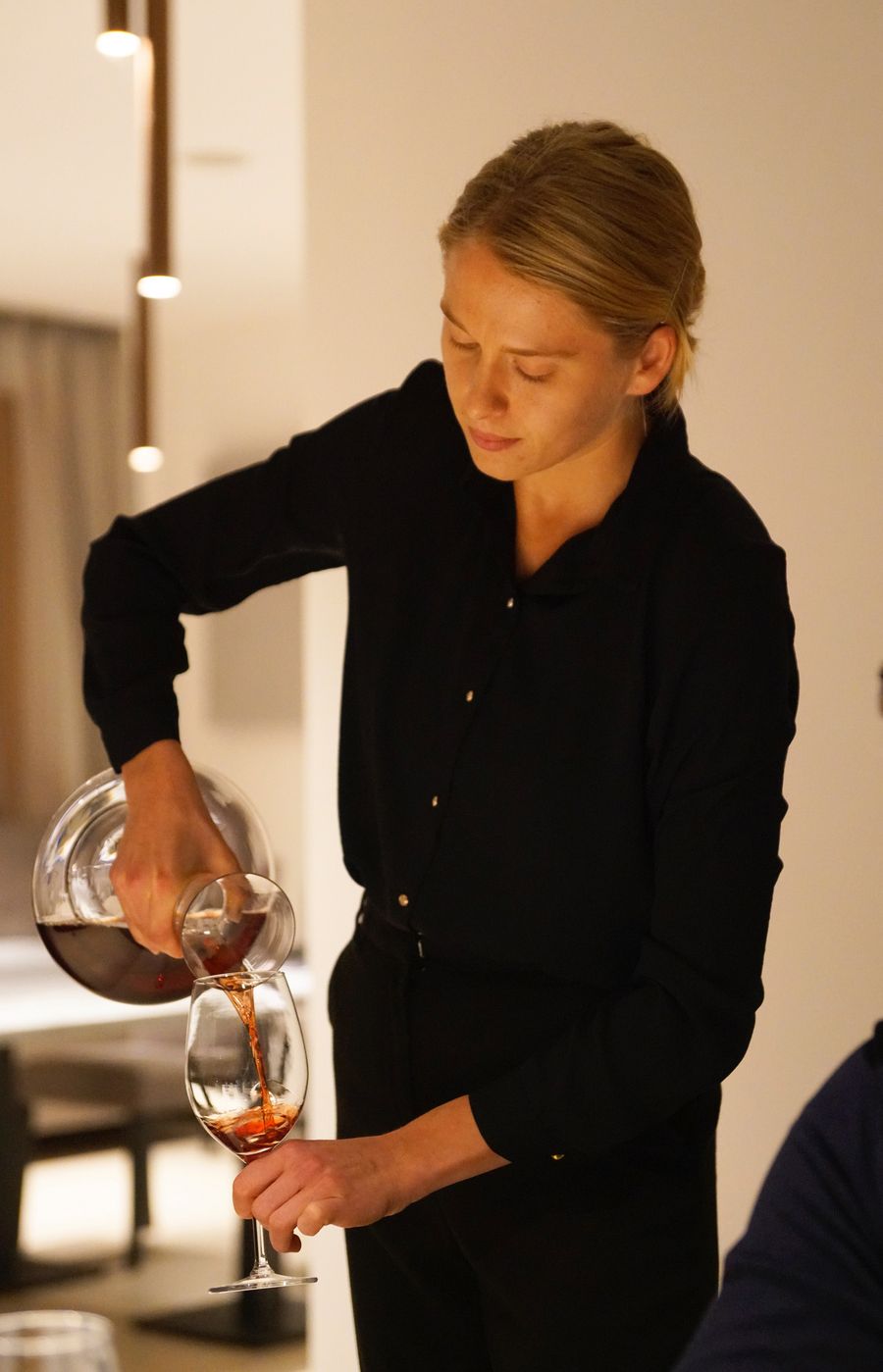 Donna che versa vino rosso in un bicchiere in un ambiente interno poco illuminato.