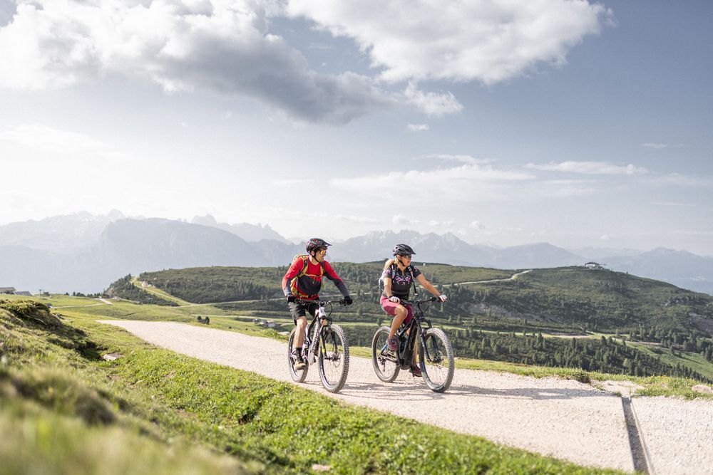 Zwei Fahrradfahrer auf einem Bergweg mit grünem Hügel- und Bergpanorama