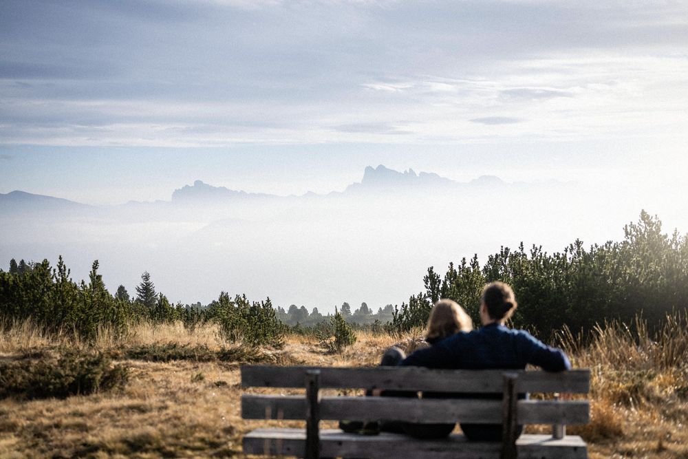 A couple sitting on a bench in a field looking at fog-covered mountains in the distance.