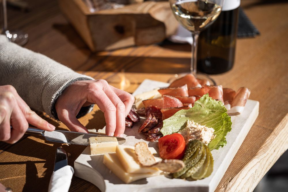 Person schneidet Käse auf einer Platte mit Wurst, Gemüse und Weißwein im Hintergrund.