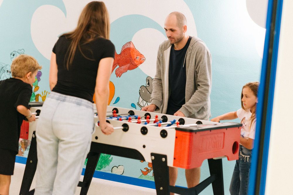 Quattro persone, due adulti e due bambini, giocano a calcio balilla in una stanza con pareti decorate con disegni marini.
