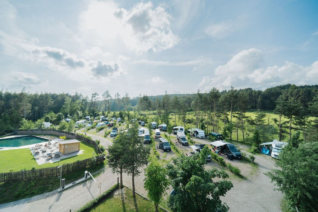 Campeggio con camper e area piscina circondata da alberi sotto un cielo parzialmente nuvoloso