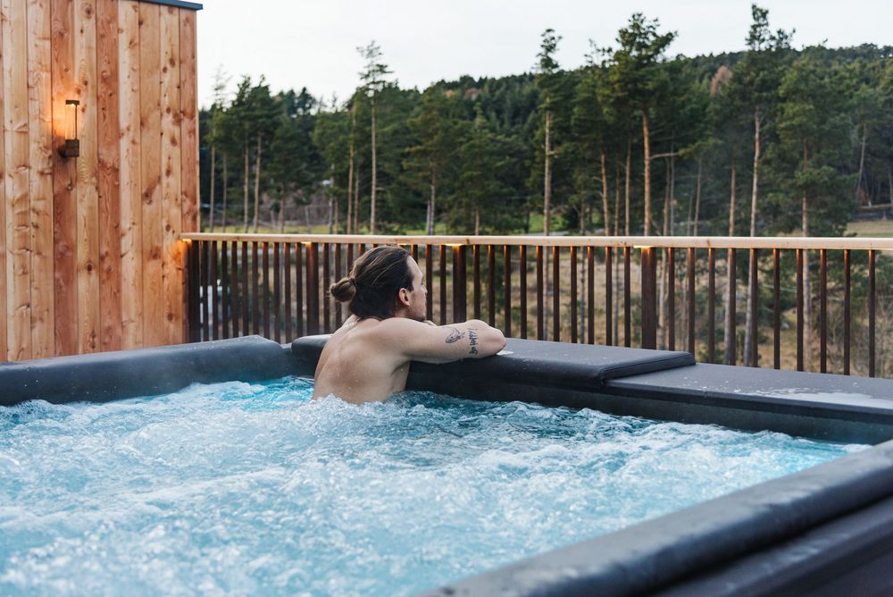 Person relaxing in a hot tub on a wooden deck overlooking trees.