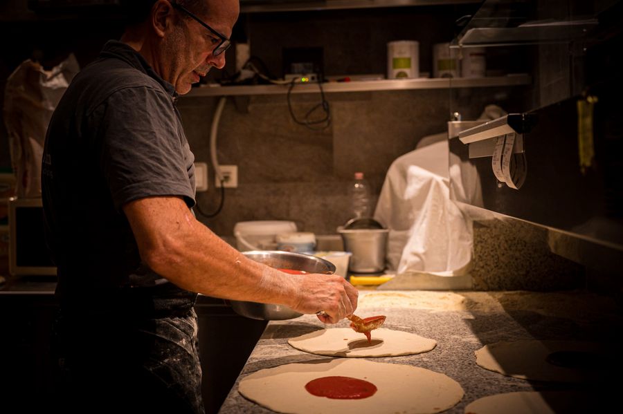 Man spreading tomato sauce on pizza dough in a kitchen.