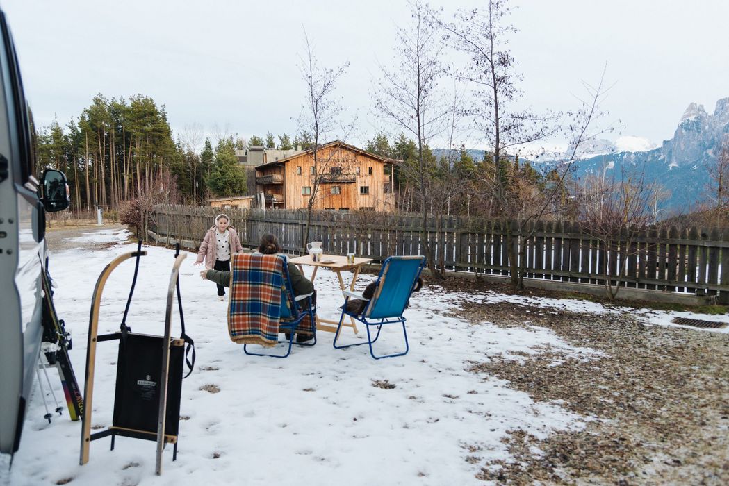 Persone sedute su sedie con una coperta durante una giornata nevosa vicino a una baita di legno e montagne sullo sfondo.