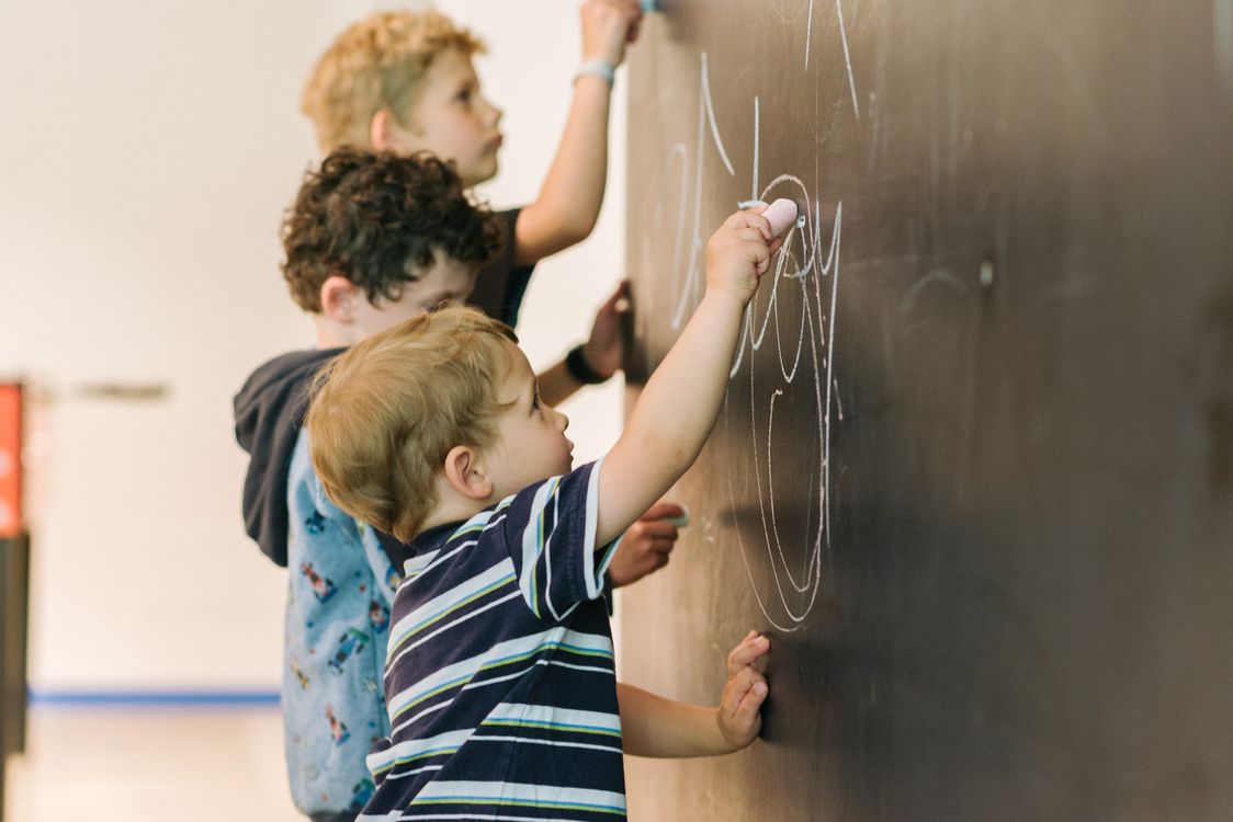 Tre bambini piccoli disegnano su una lavagna con gessetti colorati.