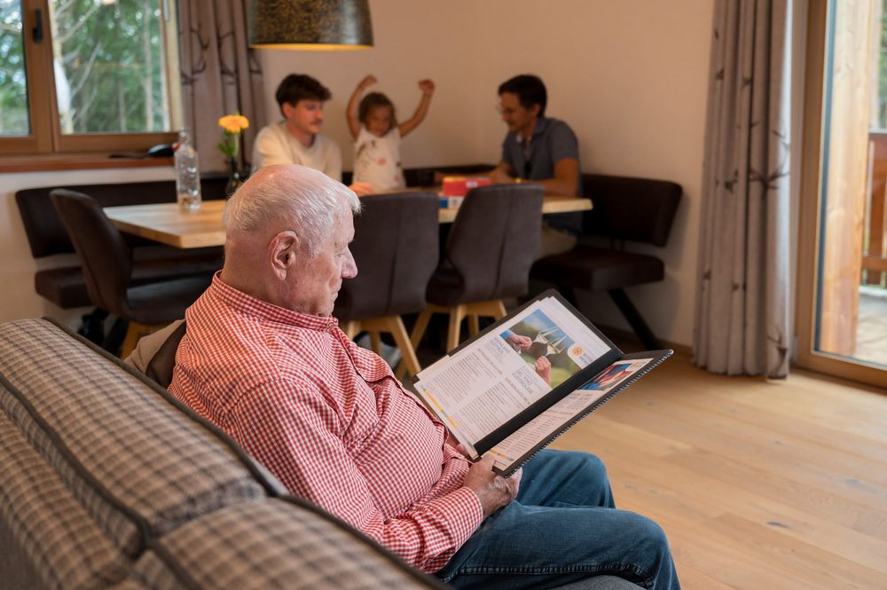 Anziano seduto sul divano che legge un libro, dietro a un tavolo due uomini e una bambina giocano e si divertono. Anziano seduto sul divano che legge un libro, dietro a un tavolo due uomini e una bambina giocano e si divertono.