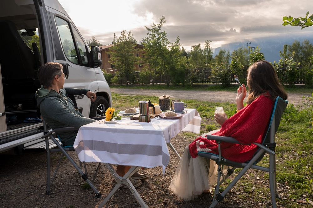 Zwei Personen sitzen an einem Campingtisch vor einem Wohnmobil und genießen eine Mahlzeit im Freien.