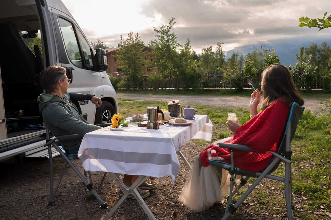 Due persone sedute all'aperto vicino a un camper con tavolo apparecchiato per colazione.