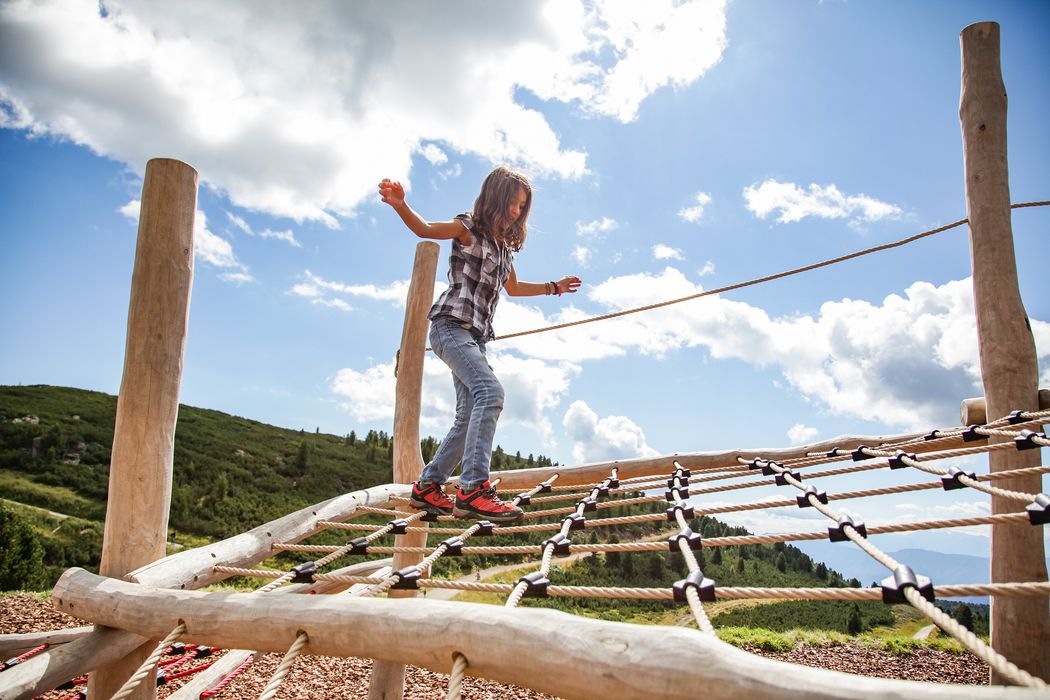 Bambina che cammina su una rete di corda in un parco avventura all'aperto sotto un cielo azzurro con nuvole.