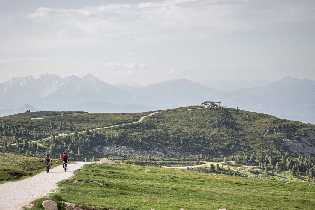 Due ciclisti su una strada di montagna con colline verdi e cielo nuvoloso sullo sfondo