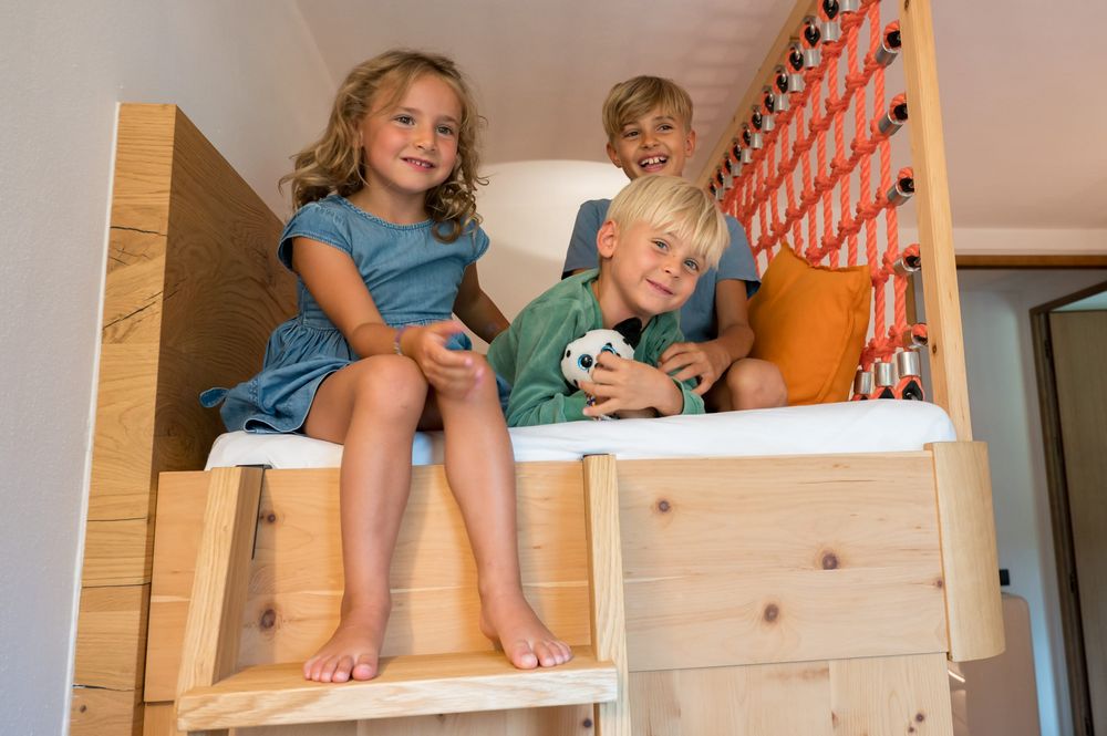Tre bambini felici seduti su un letto in legno con una rete arancione sullo sfondo.
