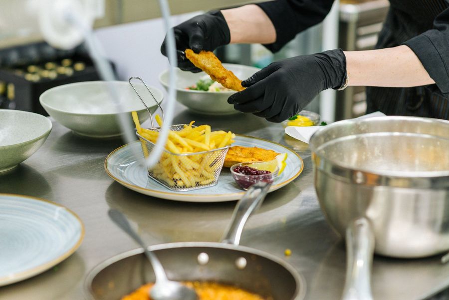 Mani con guanti neri che preparano piatto con pesce fritto, patatine e salsa