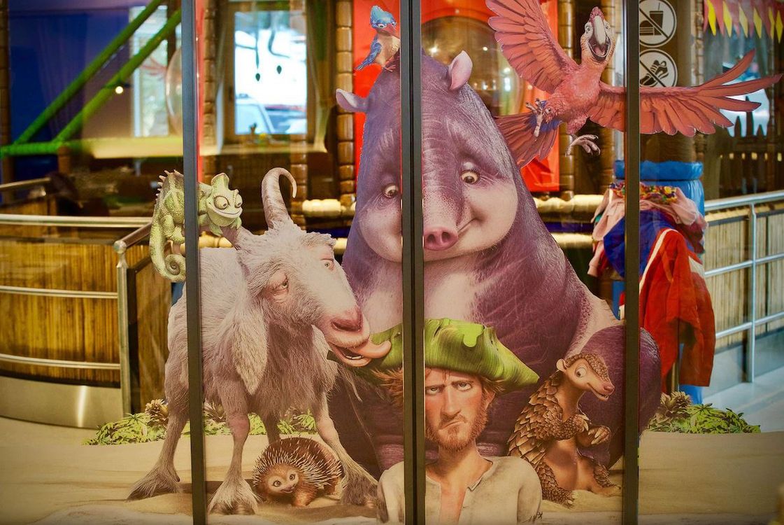 Carta da parati con personaggi animati tra cui una capra, un uomo con cappello verde, un porcospino e uccelli colorati in un ambiente interno.