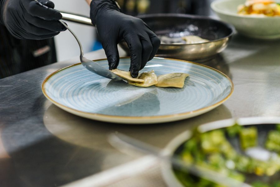 Mani con guanti neri che posizionano ravioli su un piatto blu in una cucina.