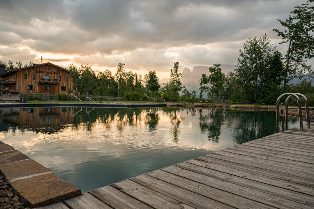 Piscina naturale con tavolato in legno, alberi e casa di montagna al tramonto