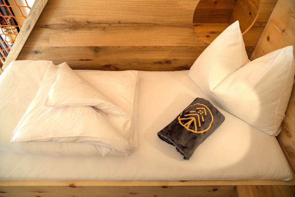 Single bed with white pillows, white sheets, and a folded gray towel with a yellow emblem on a wooden frame.