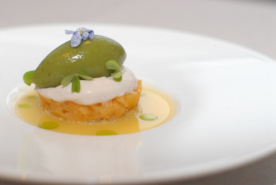 Dessert elegante con crema bianca, gelato verde, decorato con foglie verdi e fiore viola su piatto bianco.
