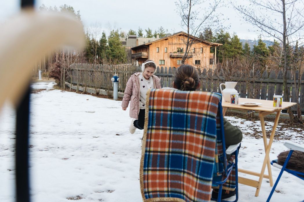 Bambina felice con cuffie paraorecchie sulla neve davanti a una persona seduta con coperta su una sedia all'aperto, tavolo con tazze e brocca