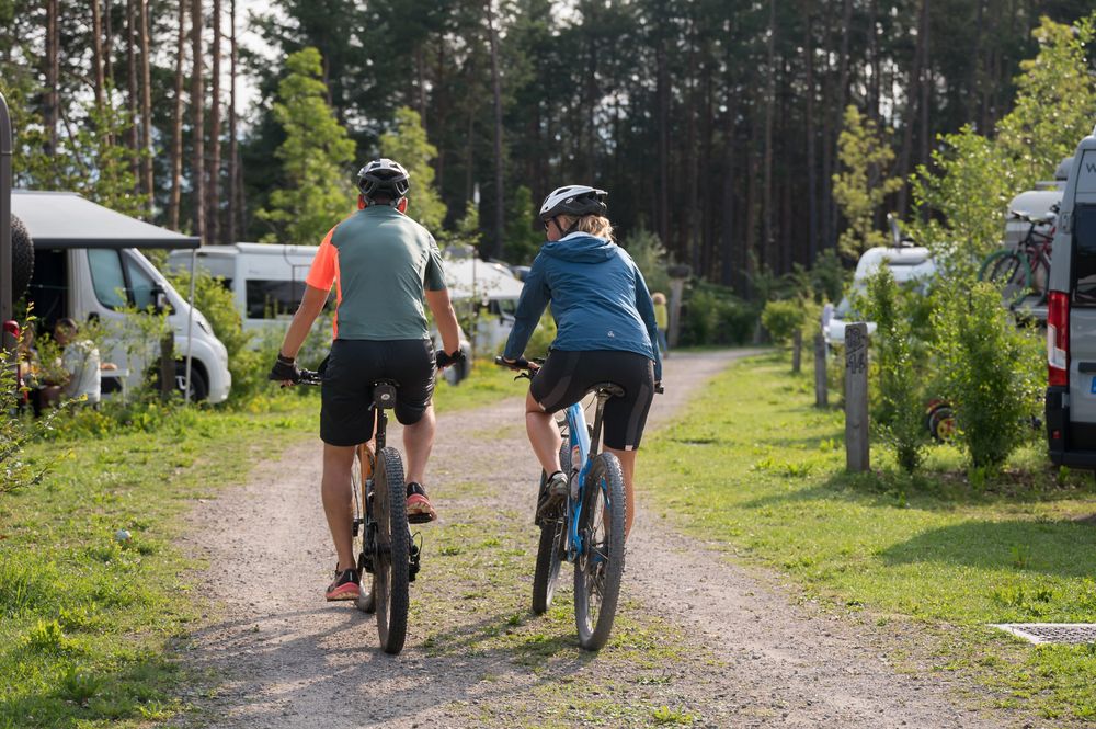 Zwei Radfahrer fahren auf einem Waldweg zwischen Wohnmobilen.