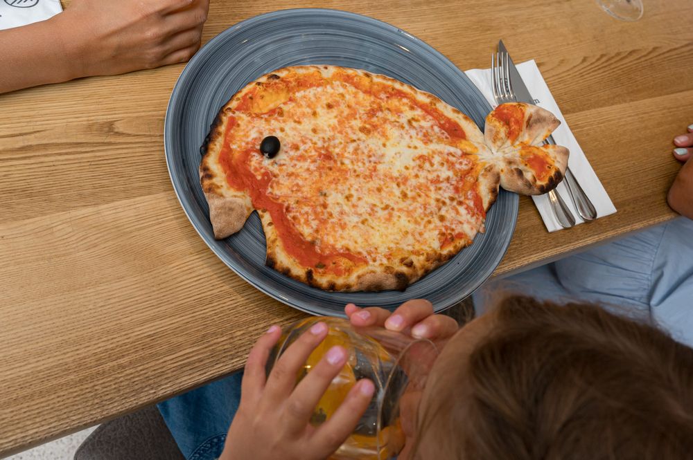 Fish-shaped cheese pizza on a plate with people around the table.