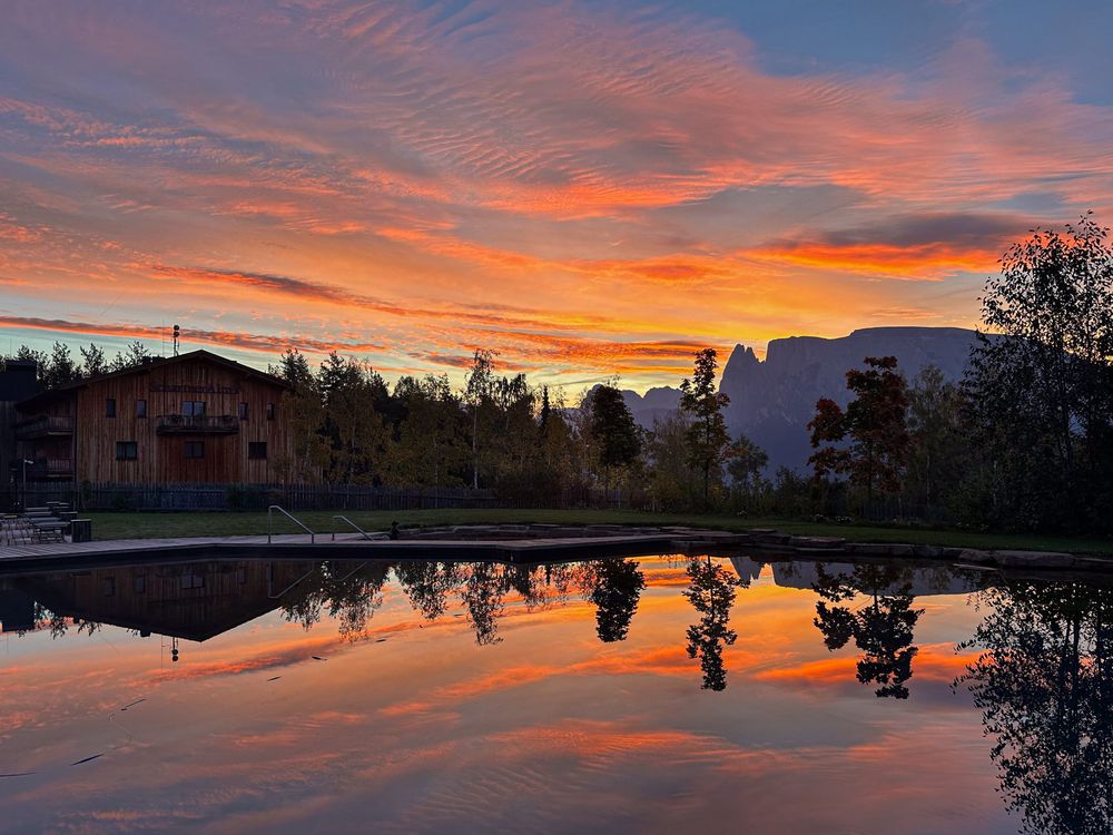 Spiegelung eines farbenfrohen Sonnenuntergangs mit Bäumen, Bergen und Gebäuden auf ruhigem Wasser