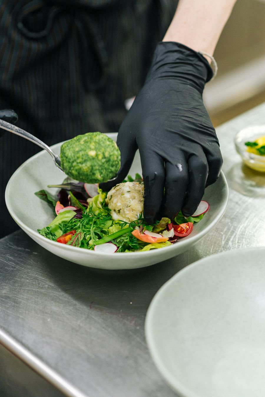 Mano con guanto nero che prepara un'insalata con verdure fresche e polpette verdi e bianche in una ciotola grigia.