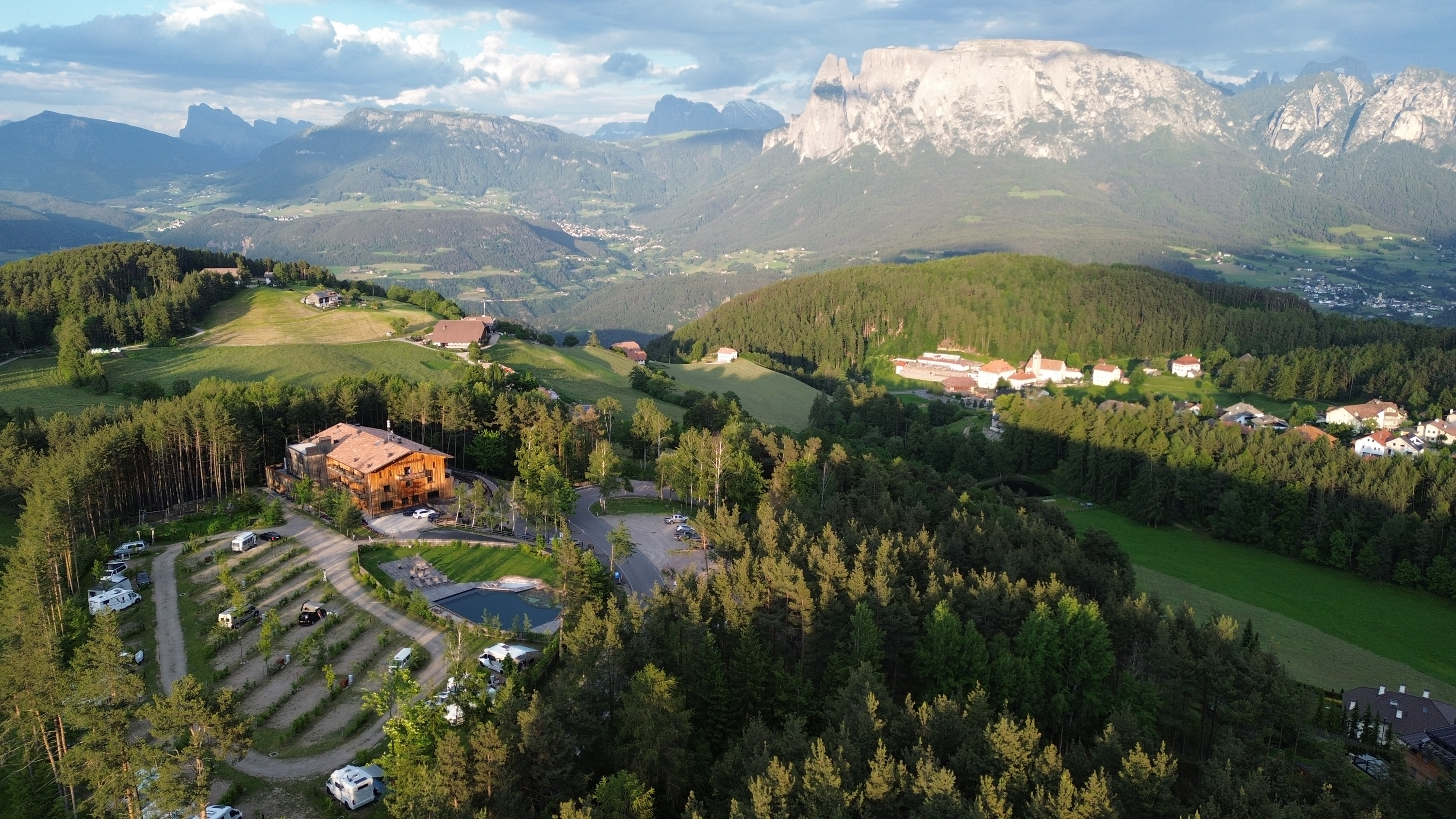 Panorama montano con alberi, prati e case in valle, cielo parzialmente nuvoloso.