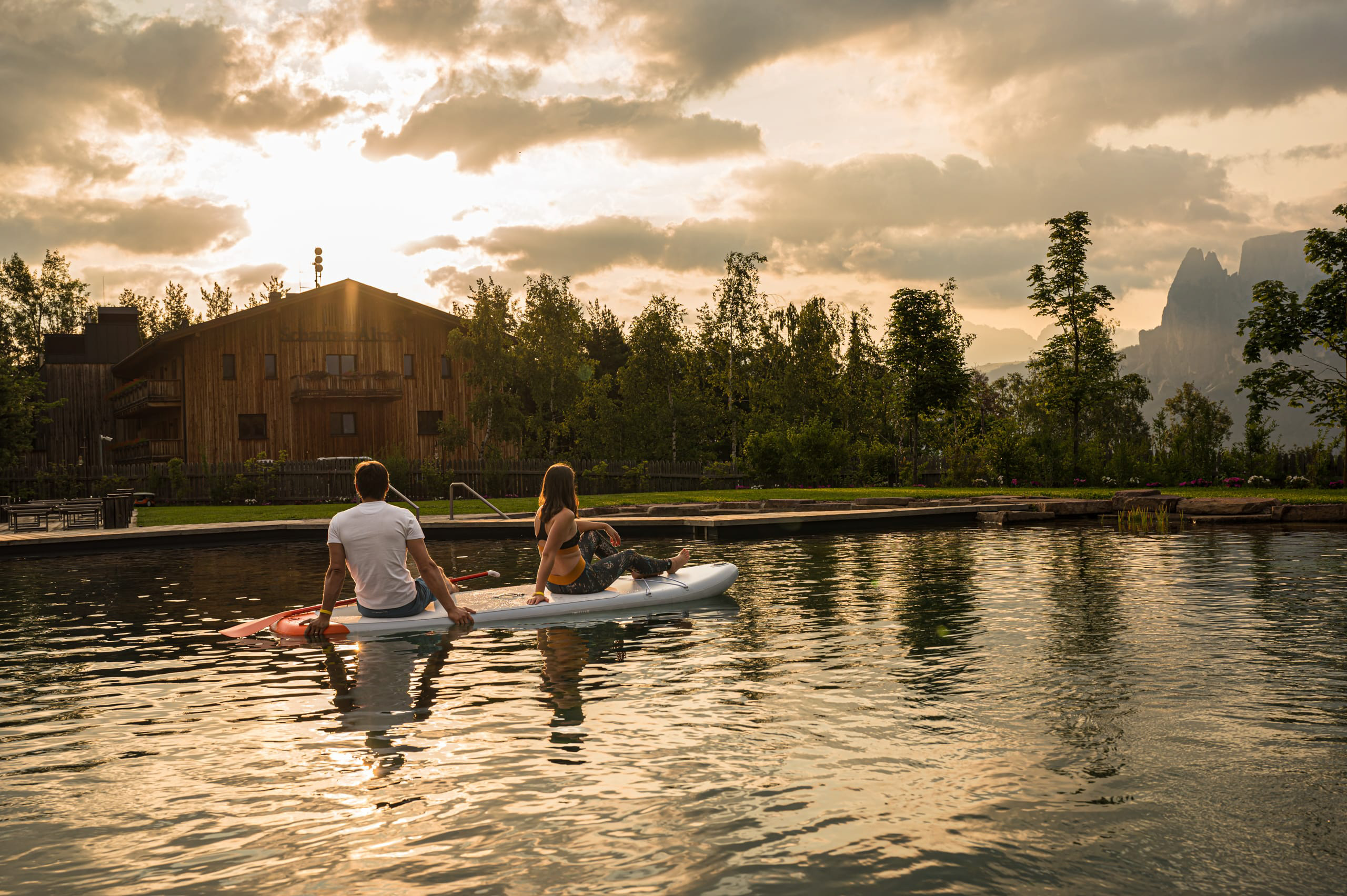 Due persone sedute su una tavola da paddle in acqua al tramonto, con una casa in legno e alberi sullo sfondo.